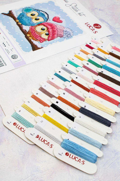 Colorful thread spools with 'LUCAS' branding on a cross-stitching background